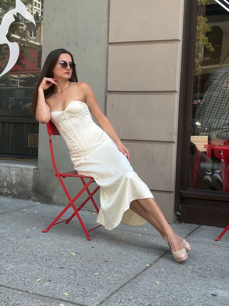 Woman in sunglasses and a pearl necklace leaning back on a red folding chair against a stone wall, in a strapless ivory corset dress whose skirt flares at the hem, with nude platform heels.