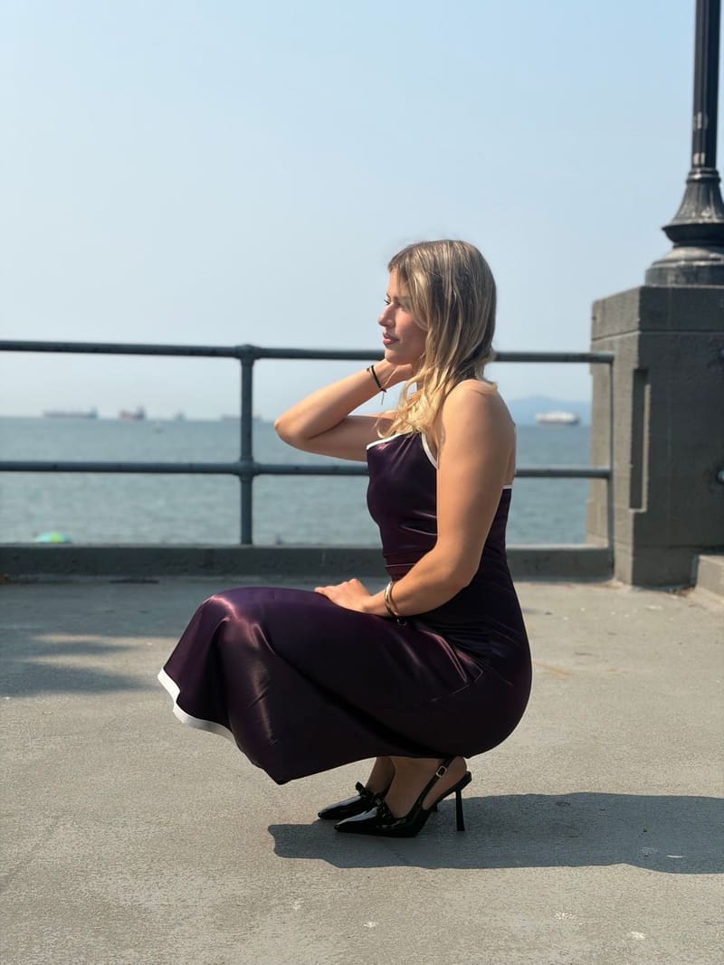 Blonde woman crouching in profile on a seaside promenade in a long plum slip dress with a white-banded hem and black slingback heels, a railing and open water behind her.