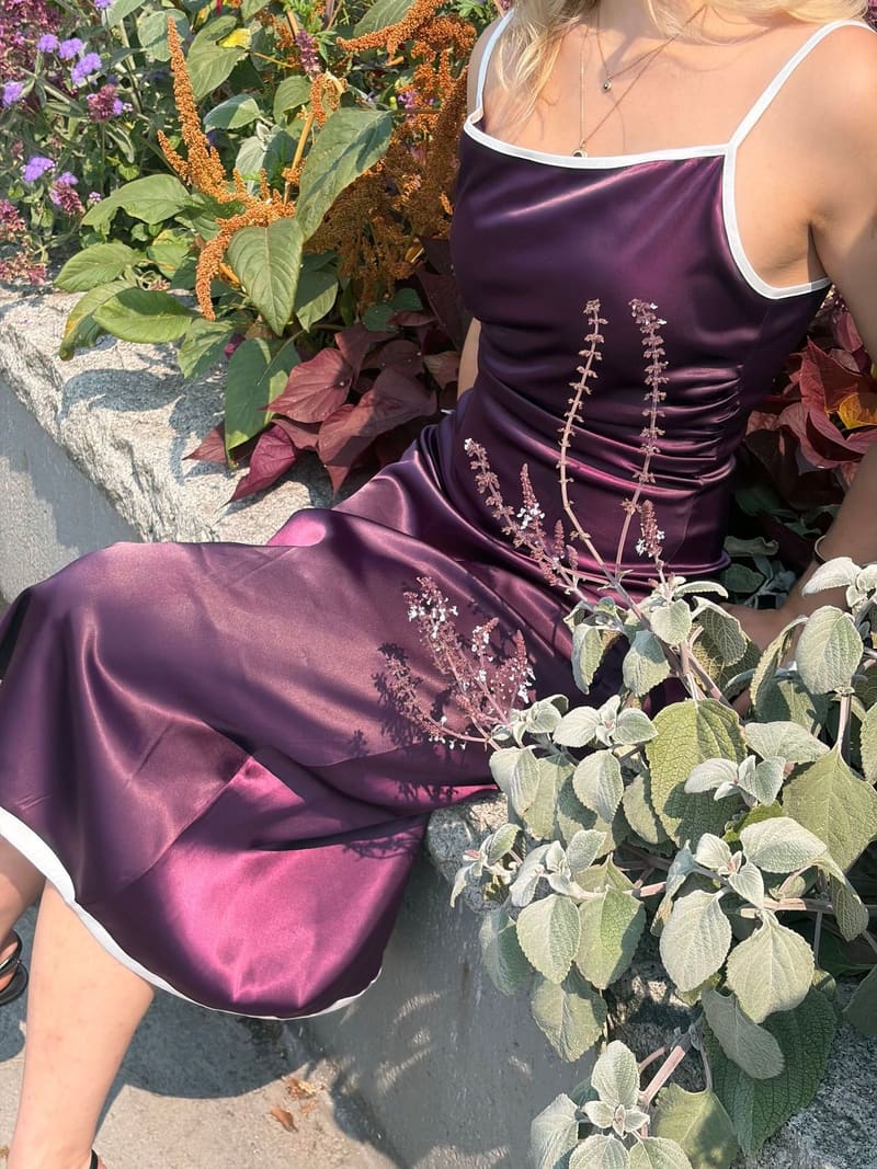 Close view of a plum slip dress on a seated woman, light running across the glossy skirt, white edging at the neckline, straps and hem, garden plants around her.