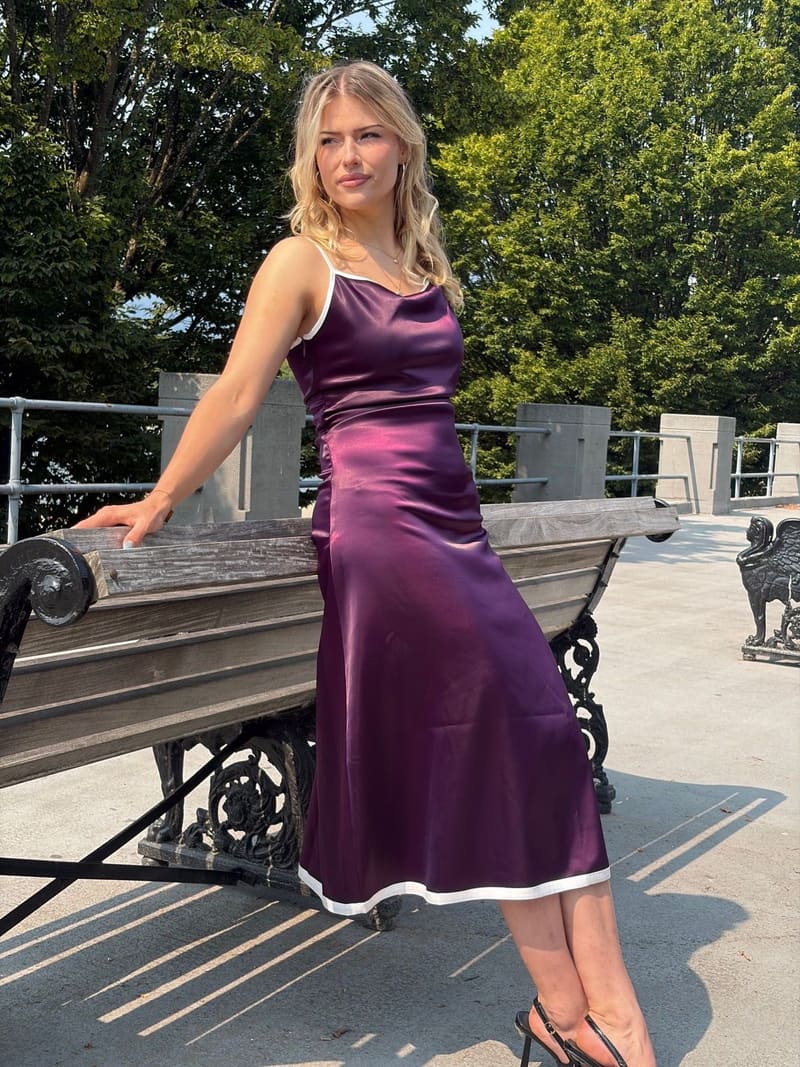 Blonde woman leaning back against a wooden bench on a sunny terrace in a long glossy plum slip dress with thin white straps, a white-edged neckline and a white band at the hem.